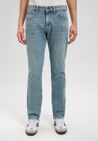 Jeans de mezclilla azul claro con un diseño ajustado, cinco bolsillos y cierre de botón. Llevados con zapatillas blancas y grises, sobre un fondo neutro.