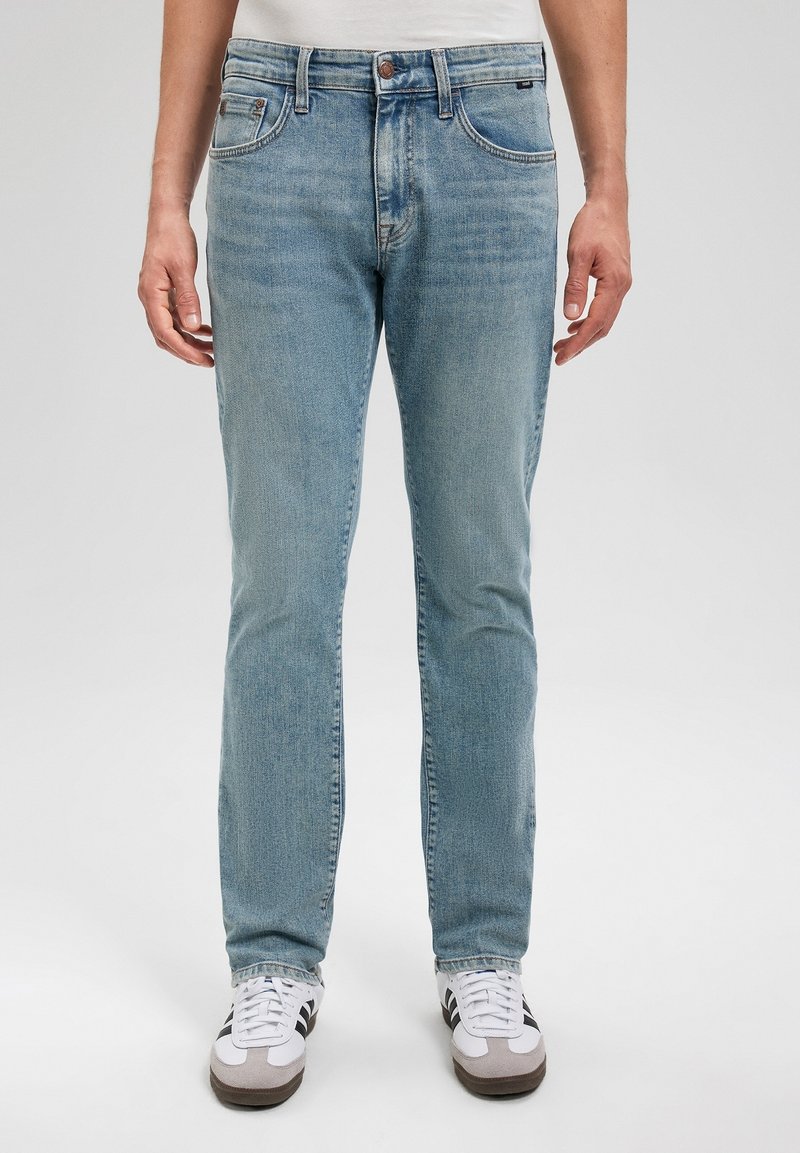 Jeans de mezclilla azul claro con un diseño ajustado, cinco bolsillos y cierre de botón. Llevados con zapatillas blancas y grises, sobre un fondo neutro.