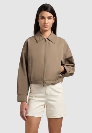 Frau trägt eine taupefarbene Jacke mit Reißverschluss und weitem Kragen sowie weiße Shorts mit hoher Taille, steht vor einem schlichten hellen Hintergrund.