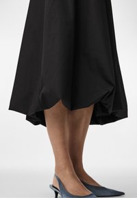 Untere Beine einer Person, die einen schwarzen Rock mit Ballonform und blaue Slingback-Kitten-Heels vor einem weißen Hintergrund trägt.