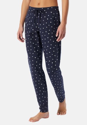 Kvinde iført marineblå pyjamasbukser med små hvide hjerte-mønstre og snøre i taljen, stående barfodet på en hvid baggrund.