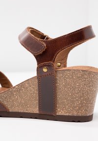 Sandalía de cuña de cuero marrón con una suela de corcho texturizada, dos correas ajustables y acentos de latón para un ajuste seguro y estilo.