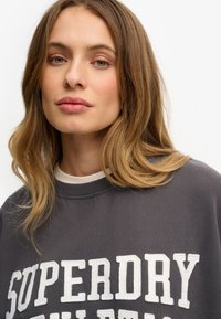 Ciemnoszara bluza z białym wyszywanym napisem "SUPERDRY". Okrągły dekolt i miękka struktura, częściowo widoczna biała koszula warstwowa pod spodem.