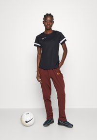 Zwart Nike sportshirt met korte mouwen en witte accenten, gecombineerd met bordeauxrode Nike broek. Witte voetbal ernaast, zwarte sportschoenen.