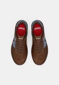 Καφέ δερμάτινα αθλητικά παπούτσια με μπλε λεπτομέρεια. Στρογγυλή μύτη, επίπεδα κορδόνια και κόκκινα πέλματα με την επιγραφή "CAMPER." Υφή στην επιφάνεια.