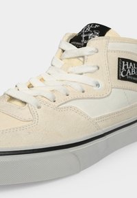 Πέδιλο Vans Half Cab ψηλού τύπου σε μπεζ και λευκό, με λευκά κορδόνια, μαύρη και λευκή ετικέτα λογότυπου στο πλάι και στη γλώσσα, και λαστιχένια σόλα.