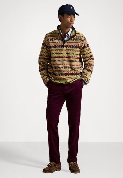 Knit pullover with a green base and colorful geometric patterns; paired with burgundy corduroy pants and brown lace-up shoes. Navy cap on head.