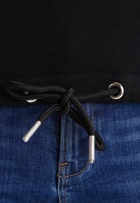 Sudadera negra con dobladillo y cordón ajustable, presenta ojales y extremos de metal plateado, combinada con jeans de mezclilla azules.