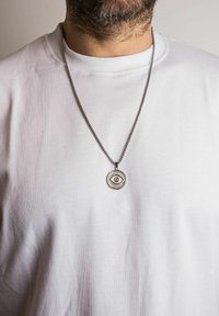 Sølv kædedesign med en rund vedhæng med et øje, båret over en enkel hvid t-shirt. Tekstureret metallisk overflade med detaljerede accenter.