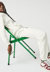 Vit hoodie och joggers med ribbad textur, sittande på en grön vikstol. Skorna har en burgundy och vit design med rutiga detaljer.