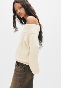 Maglione a spalle scoperte crema con colletto e polsini a coste, abbinato a jeans in denim scuro. Tessuto morbido; vestibilità rilassata; maniche lunghe in vista.