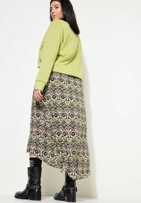 Groene sweatshirt overlay, maxi rok met patronen van bloemen en geometrische motieven in groen, roze en zwart, gecombineerd met kniehoge zwarte laarzen.