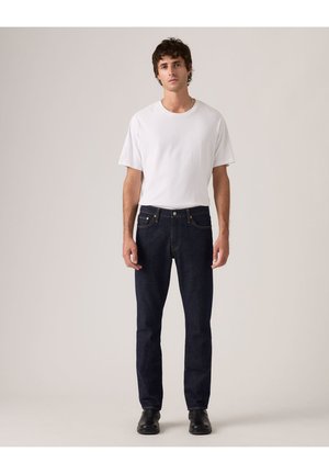 Joven de pie con camiseta blanca lisa, vaqueros azul oscuro y zapatos negros sobre un fondo neutro claro.