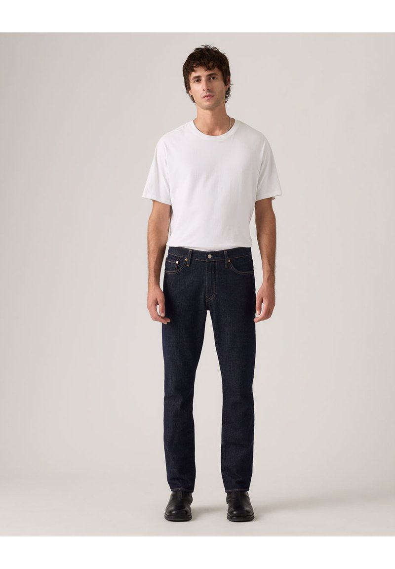 Giovane uomo in piedi che indossa una maglietta bianca semplice, jeans blu scuro e scarpe nere su uno sfondo chiaro e neutro.