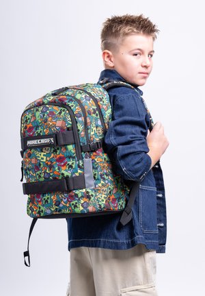 Junge mit Jeansjacke und hellen Hosen trägt einen bunten Minecraft-Rucksack mit mehreren Fächern und schwarzen Trägern.
