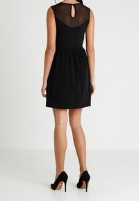 Robe noire avec un dos en tulle transparent, sans manches, taille froncée et détail d'encolure festonnée. Portée avec des escarpins noirs.