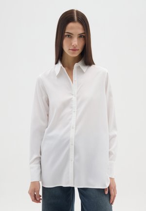 Frau mit glatten braunen Haaren, die ein lockeres weißes Hemd mit Knöpfen und dunkelblaue Jeans vor einem einfachen weißen Hintergrund trägt.