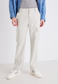Pantalones cargo beige con múltiples bolsillos, hechos de una tela ligera, con un corte de pierna recta. Combinados con zapatillas blancas y una camisa gris.
