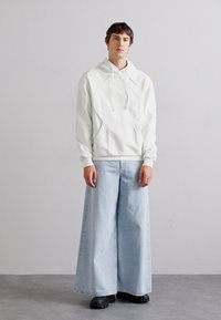 Sudadera blanca con capucha y detalles de costura, combinada con pantalones de mezclilla de pierna ancha en azul claro. Calzado negro completa el conjunto.