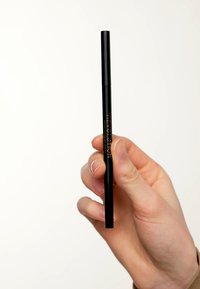 Zwarte eyelinerpen vastgehouden door een hand, met een gladde cilindrische vorm en merktext in brons. Matte afwerking en een afgedekte punt.