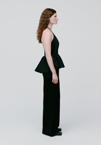 Femme aux cheveux bouclés portant un haut peplum noir à col halter, un pantalon à jambes larges et des boucles d'oreilles anneaux, debout de profil sur un fond uni.