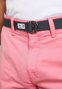 Rosa bomullsshorts med en slät textur, försedd med ett svart vävt bälte som är säkrat med en metallring, synliga sömnadsdetaljer och en etikett.