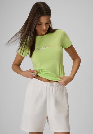 Kobieta z długimi brązowymi włosami, ubrana w jasnografitowy T-shirt "Tropical" i białe szorty, lekko podnosząca koszulkę, aby odsłonić talię.