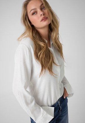 Junge Frau mit langen blonden Haaren trägt ein weißes Hemd mit Knöpfen und blaue Jeans, posiert mit einer Hand in der Tasche vor einem schlichten Hintergrund.