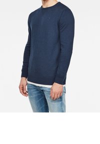 Námořnický modrý pletený svetr s kulatým výstřihem, žebrovanými manžetami a zakončením. Obsahuje malé logo na hrudi a je vrstvený přes bílou košili.