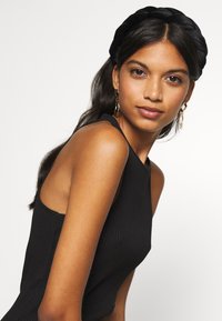 Μαύρη ραβδωτή αμάνικη μπλούζα με φαρδιές τιράντες, που διαθέτει ψηλό λαιμό. Το μοντέλο φοράει πλεξούδα μαύρη κορδέλα για τα μαλλιά και χρυσά σκουλαρίκια κρίκους.