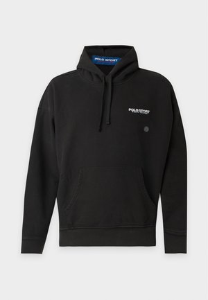 Homme portant un sweat à capuche Polo Sport noir, pantalon blanc et mocassins beiges, debout sur un fond blanc uni.
