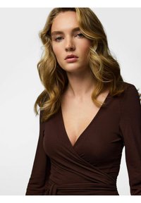 Brūns pārsienams tops ar gariem piedurknēm un dziļu V-veida izgriezumu. Audums ir mīksts un gluds, ar jostu viduklī.