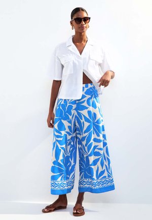 Weißes kurzärmeliges Hemd; weit geschnittene Hose mit blau-weißem Blattmuster und Randdesign; kombiniert mit Sonnenbrille und Sandalen.