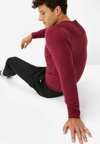Burgundroter Strickpullover mit gerippten Akzenten an den Ärmeln, kombiniert mit schwarzen Hosen. Das Model sitzt und präsentiert die Passform und Textur des Pullovers.