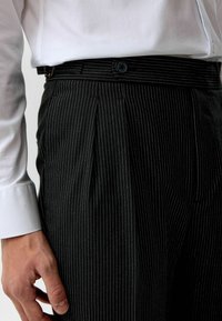 Zwarte krijtstreep broeken met een geplooid ontwerp, voorzien van een zijknop en een textuurstof. Draagt een wit overhemd met lange mouwen.