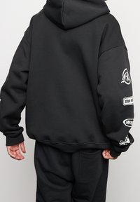 Sweat à capuche noir en tissu doux avec une grande capuche, présentant des impressions de logo sur les manches ainsi que des poignets et un ourlet côtelés.