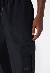 Primer plano de la mano de una persona descansando cerca de la solapa del bolsillo de unos pantalones cargo negros con detalles de costura y una pequeña etiqueta de tela.