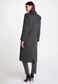 Abrigo largo de lana gris con cuello con muesca, costuras verticales y una abertura en la parte trasera. Llevado sobre jeans grises y zapatos de tacón altos negros.