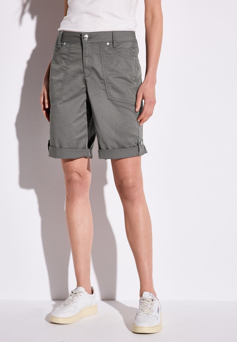 Un pantalon court gris en tissu léger, doté de revers roulés, de poches avant et de détails en boutons. Associé à des baskets blanches.