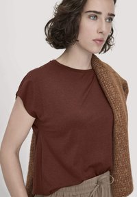 Braunes kurzärmliges T-Shirt aus weichem Stoff mit lässigem Schnitt, kombiniert mit einer übergroßen gestrickten beige Farbkarte. Der Hintergrund ist neutral.