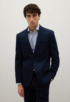 Blazer azul marino con dos botones, solapas de muesca y bolsillos laterales, combinado con una camisa de botones a rayas. Tejido suave, ajuste entallado.