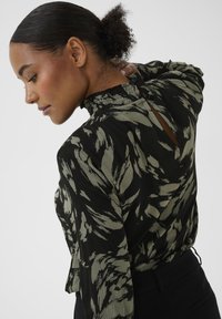 Zwarte blouse met een abstract olijfgroen bladpatroon, gedetailleerde halslijn met plooien en knoopsluiting aan de achterkant. Zachte, gestructureerde stof.