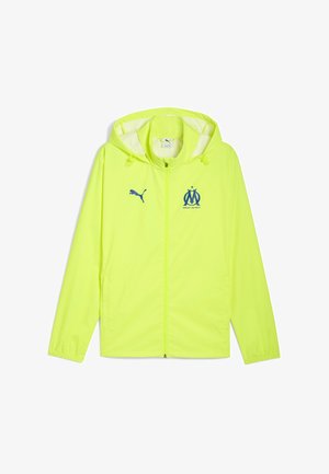 Giacca leggera verde lime con cappuccio, con logo Puma blu e stemma di Marsiglia sul petto. Chiusura frontale con zip e polsini elasticizzati.