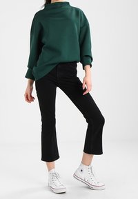 Persoon in een donkergrijze oversized trui, zwarte cropped jeans en witte hoge sneakers, staande met één been iets opgetild.