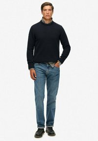 Pull marine avec col haut, superposé à une chemise à carreaux, associé à un jean bleu et des chaussures noires. Présente une coupe ajustée.