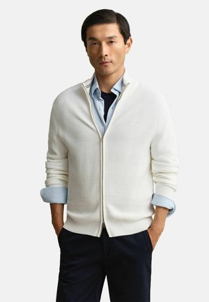 Mann trägt weißen Reißverschluss-Pullover über hellblauem Hemd mit hochgekrempelten Ärmeln und dunkler Hose, Hände in den Taschen, neutraler Hintergrund.