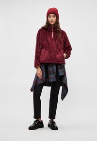 Bordowy polar z połowicznym zamkiem, z podszewką w kieszeni, noszony na niebiesko-zielonej koszuli w kratę zawiązanej w talii, w połączeniu z czarnymi dżinsami i błyszczącymi butami.