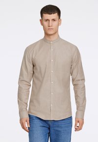 Camisa de manga larga, con cuello, en tejido beige claro con una textura sutil, que presenta un cierre de botones en el frente y en los puños, combinada con jeans azules.