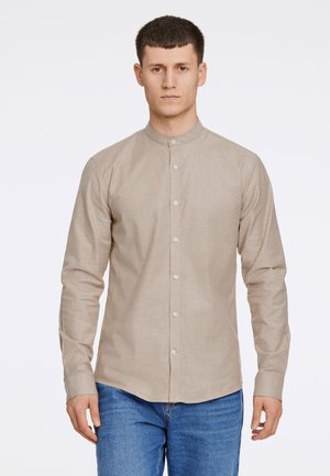 Camisa de manga larga, con cuello, en tejido beige claro con una textura sutil, que presenta un cierre de botones en el frente y en los puños, combinada con jeans azules.