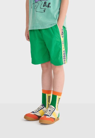 Enfant portant un short vert avec des bandes latérales jaunes, des chaussettes hautes colorées avec du texte, et des baskets multicolores, debout devant un fond uni.